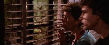 Movie still from “The Lost Boys” (1987), directed by Joel Schumacher – A young man looking out of a window through a wooden slat blind; Close Up shot, Low angle