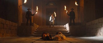 Movie still from “The Lost City” (2022), directed by Aaron Nee – A man sitting on the steps of a building holding torches; Wide shot, Low angle