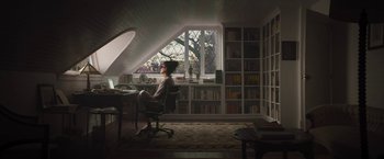 Movie still from “The Lost City” (2022), directed by Aaron Nee – A woman sitting at a computer desk in front of a window; Wide shot, High angle