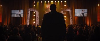 Movie still from “The Lost City” (2022), directed by Aaron Nee – A man standing in front of an audience in front of lights; Wide shot, Over the shoulder angle