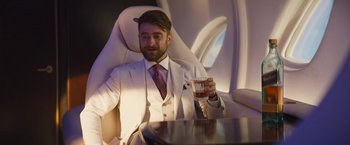Movie still from “The Lost City” (2022), directed by Aaron Nee – A man in a white suit sitting at a table with a glass of whiskey; Medium shot, Over the shoulder angle