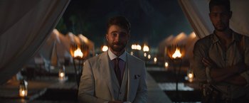 Movie still from “The Lost City” (2022), directed by Aaron Nee – A man wearing a suit and tie standing in front of a fire pit; Medium shot, Over the shoulder angle