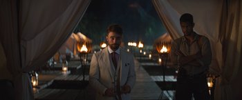 Movie still from “The Lost City” (2022), directed by Aaron Nee – A man in a white suit standing in front of lit torches at night; Wide shot, Over the shoulder angle