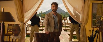 Movie still from “The Lost City” (2022), directed by Aaron Nee – A man in a brown suit standing next to a brown chair; Medium shot, Over the shoulder angle