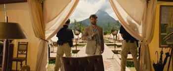 Movie still from “The Lost City” (2022), directed by Aaron Nee – A man in a suit standing next to a chair in front of a mountain; Wide shot, Over the shoulder angle