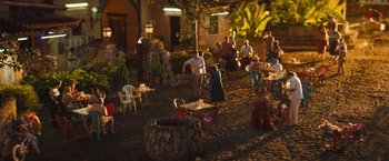 Movie still from “The Lost City” (2022), directed by Aaron Nee – A group of people sitting at tables outside at night; Extreme Wide shot, High angle