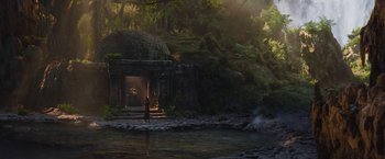 Movie still from “The Lost City” (2022), directed by Aaron Nee – A person standing near a body of water near a cave; Extreme Wide shot, High angle