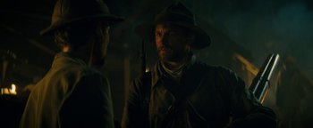 Movie still from “The Lost City of Z” (2016), directed by James Gray – A man in a hat holding a rifle in a dark room; Close Up shot, Over the shoulder angle