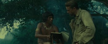 Movie still from “The Lost City of Z” (2016), directed by James Gray – A man is reading a book to another man in the woods; Medium shot, Over the shoulder angle