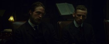 Movie still from “The Lost City of Z” (2016), directed by James Gray – Two men in suits sitting next to each other in a dark room; Medium shot, Over the shoulder angle