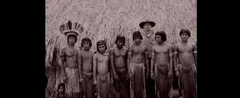 Movie still from “The Lost City of Z” (2016), directed by James Gray – A group of people standing next to each other in front of a thatched hut; Wide shot, High angle