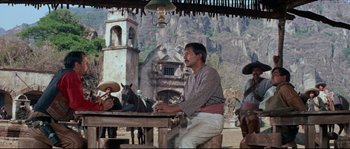 Movie still from “The Magnificent Seven” (1960), directed by John Sturges – A man sitting at a table in front of a building; Wide shot, Over the shoulder angle