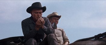 Movie still from “The Magnificent Seven” (1960), directed by John Sturges – Two men in cowboy hats are sitting on a bench; Medium shot, Low angle