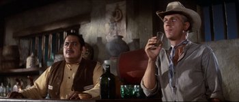 Movie still from “The Magnificent Seven” (1960), directed by John Sturges – Two men sitting at a table with a drink; Medium shot, Low angle