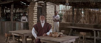 Movie still from “The Magnificent Seven” (1960), directed by John Sturges – A man sitting at a table in front of a brick building; Wide shot, Over the shoulder angle