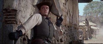 Movie still from “The Magnificent Seven” (1960), directed by John Sturges – A man wearing a cowboy hat and a vest leaning against a stone wall; Medium shot, Low angle