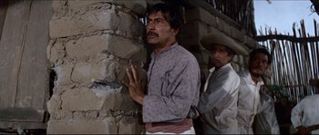 Movie still from “The Magnificent Seven” (1960), directed by John Sturges – A man standing next to a stone wall with his hands clasped; Medium shot, Low angle