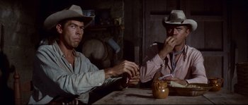Movie still from “The Magnificent Seven” (1960), directed by John Sturges – Two men sitting at a wooden table eating food; Medium shot, Over the shoulder angle