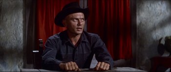 Movie still from “The Magnificent Seven” (1960), directed by John Sturges – A man wearing a cowboy hat sitting at a table in front of a red curtain; Medium shot, Low angle