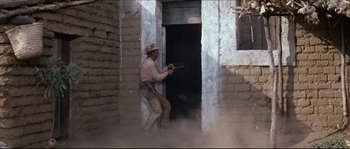 Movie still from “The Magnificent Seven” (1960), directed by John Sturges – A man holding a gun while standing in front of a building; Wide shot, Over the shoulder angle