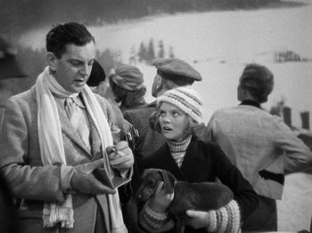 Movie still from “The Man Who Knew Too Much” (1934), directed by Alfred Hitchcock – A man and a woman standing next to each other holding a dog; Medium shot, Over the shoulder angle