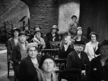 Movie still from “The Man Who Knew Too Much” (1934), directed by Alfred Hitchcock – A group of people sitting in a room with a brick wall behind them; Wide shot, High angle