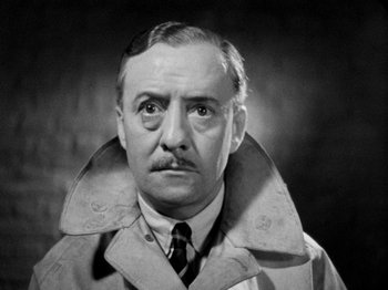 Movie still from “The Man Who Knew Too Much” (1934), directed by Alfred Hitchcock – An older man wearing a trench coat and tie; Close Up shot, Low angle