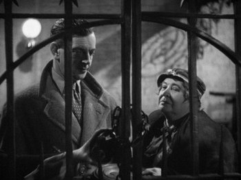 Movie still from “The Man Who Knew Too Much” (1934), directed by Alfred Hitchcock – A man and a woman are standing next to each other in front of a metal fence; Medium shot, Over the shoulder angle