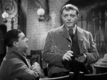 Movie still from “The Man Who Knew Too Much” (1934), directed by Alfred Hitchcock – A man smoking a cigarette while talking to another man in front of him; Medium shot, Over the shoulder angle
