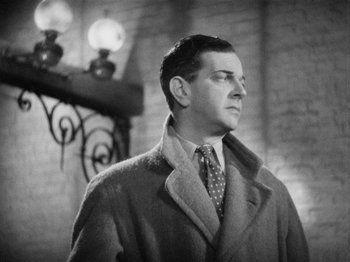 Movie still from “The Man Who Knew Too Much” (1934), directed by Alfred Hitchcock – A man wearing a coat and tie standing in front of a brick wall; Close Up shot, Low angle