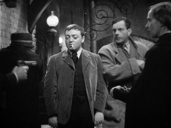 Movie still from “The Man Who Knew Too Much” (1934), directed by Alfred Hitchcock – A black - and - white photo of a man smoking a cigarette; Medium shot, Over the shoulder angle