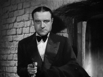 Movie still from “The Man Who Knew Too Much” (1934), directed by Alfred Hitchcock – A man in a suit and tie holding a gun; Close Up shot, Low angle