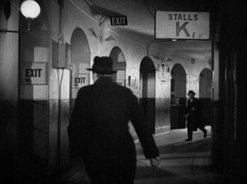 Movie still from “The Man Who Knew Too Much” (1934), directed by Alfred Hitchcock – A black and white photo of a man in a hat walking down a street; Wide shot, Over the shoulder angle