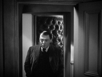 Movie still from “The Man Who Knew Too Much” (1934), directed by Alfred Hitchcock – A black and white photo of a man standing in a doorway; Medium shot, High angle