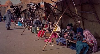 Movie still from “The Man Who Knew Too Much” (1956), directed by Alfred Hitchcock – A group of people sitting on a street under a tent; Wide shot, High angle