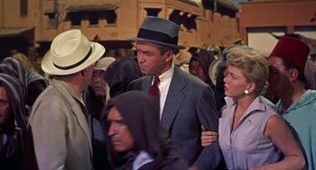 Movie still from “The Man Who Knew Too Much” (1956), directed by Alfred Hitchcock – A man in a suit and a hat standing next to a crowd of people; Medium shot, Over the shoulder angle