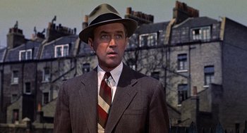 Movie still from “The Man Who Knew Too Much” (1956), directed by Alfred Hitchcock – A man in a suit and hat standing in front of a row of houses; Close Up shot, Low angle