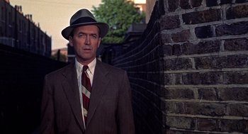 Movie still from “The Man Who Knew Too Much” (1956), directed by Alfred Hitchcock – A man in a suit and hat standing in front of a brick wall; Close Up shot, Low angle