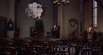 Movie still from “The Man Who Knew Too Much” (1956), directed by Alfred Hitchcock – A man and a woman are sitting at a podium in front of chairs; Wide shot, High angle