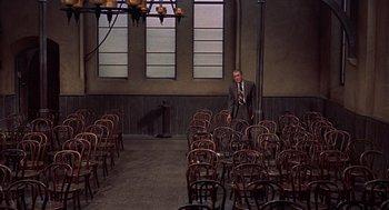 Movie still from “The Man Who Knew Too Much” (1956), directed by Alfred Hitchcock – A man in a suit and tie standing next to a bunch of wooden chairs; Wide shot, High angle