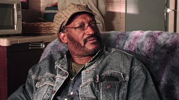 Movie still from “The Man from Earth” (2007), directed by Richard Schenkman – An older man sitting in a chair wearing glasses and a hat; Close Up shot, Over the shoulder angle