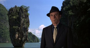 Movie still from “The Man with the Golden Gun” (1974), directed by Guy Hamilton – An older man in a suit and hat standing in front of a body of water; Medium shot, Low angle