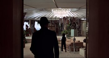 Movie still from “The Man with the Golden Gun” (1974), directed by Guy Hamilton – A man in a suit and hat in a room with furniture; Extreme Wide shot, High angle