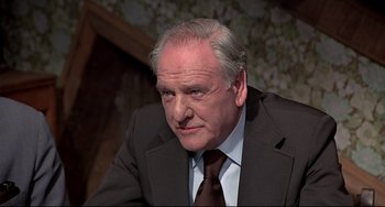 Movie still from “The Man with the Golden Gun” (1974), directed by Guy Hamilton – An older man wearing a suit and tie; Close Up shot, Over the shoulder angle