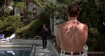 Movie still from “The Man with the Golden Gun” (1974), directed by Guy Hamilton – A man standing next to a swimming pool while another man watches; Wide shot, Over the shoulder angle