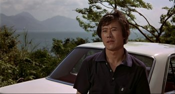 Movie still from “The Man with the Golden Gun” (1974), directed by Guy Hamilton – A man standing in front of a car on a road; Close Up shot, Low angle