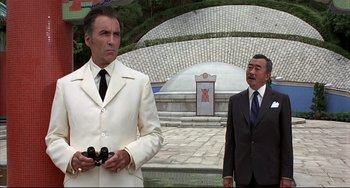 Movie still from “The Man with the Golden Gun” (1974), directed by Guy Hamilton – A man in a suit and tie standing next to another man in a suit and tie holding binoculars; Medium shot, Low angle
