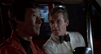 Movie still from “The Man with the Golden Gun” (1974), directed by Guy Hamilton – A man in a suit sitting next to another man in a car; Close Up shot, Over the shoulder angle