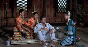 Movie still from “The Man with the Golden Gun” (1974), directed by Guy Hamilton – A man sitting on the ground next to a group of women; Wide shot, High angle