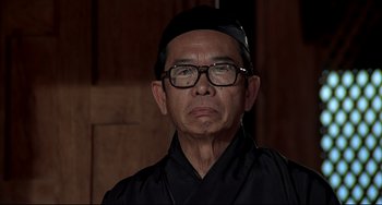 Movie still from “The Man with the Golden Gun” (1974), directed by Guy Hamilton – An older man wearing glasses and wearing a black hat; Close Up shot, Low angle